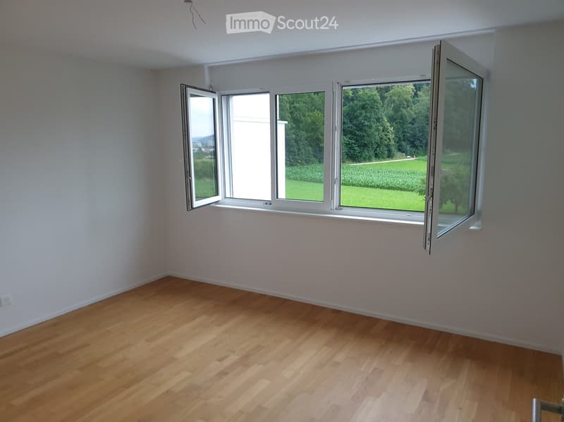 Empty room with light wooden flooring and large windows offering a view of a green landscape.