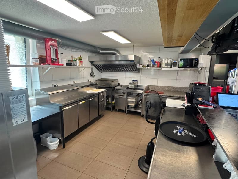 Commercial kitchen with stainless steel appliances, including a refrigerator, oven, microwave, and fan. Shelves hold various bottles and containers. Counter space is visible with a screen and other small items.