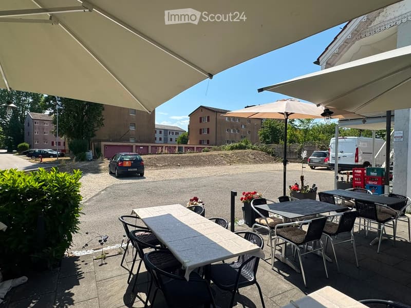 Outdoor seating area with tables, chairs, and an umbrella, surrounded by greenery and parked cars.