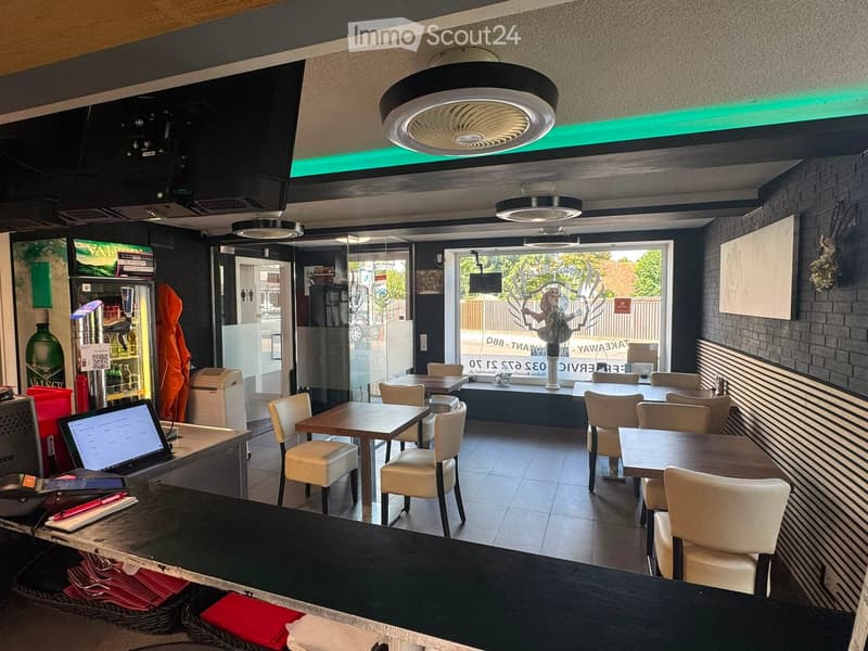 Modern restaurant interior with tables, chairs, a laptop on the counter, a refrigerator, and a fan on the ceiling.