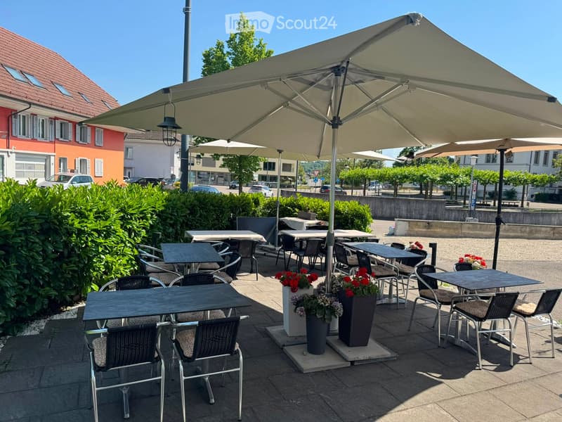 Outdoor seating area with tables, chairs, and large umbrellas, surrounded by greenery and buildings.