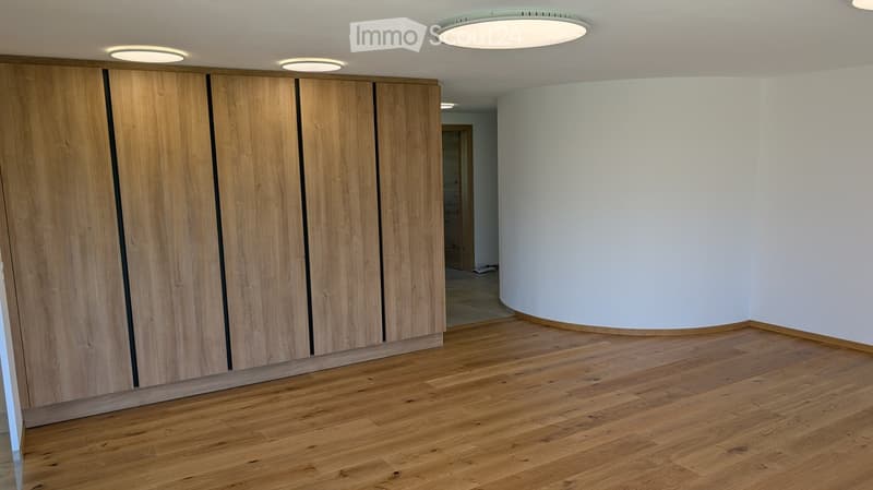 Empty room with wooden flooring, white walls, and recessed lighting. Features a large wooden wardrobe on one side and a doorway leading to another space.