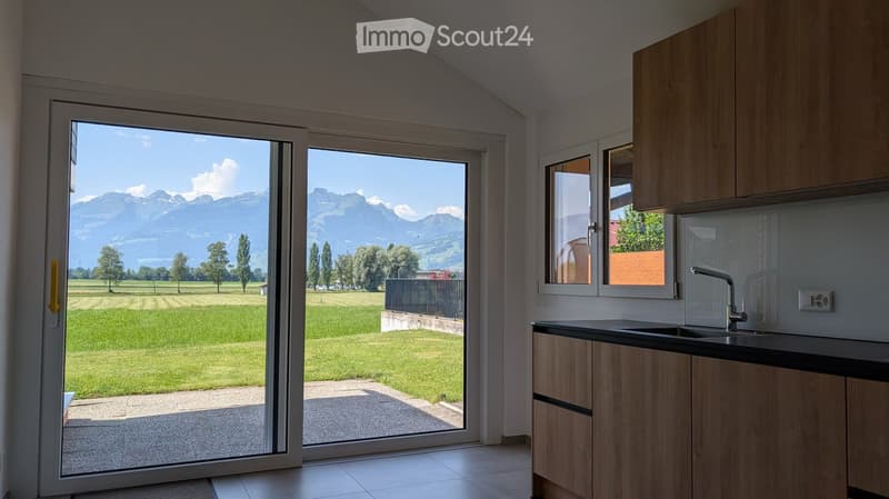 Modern kitchen with light wooden cabinets, black countertop, and a large sliding glass door opening to a scenic mountain view.