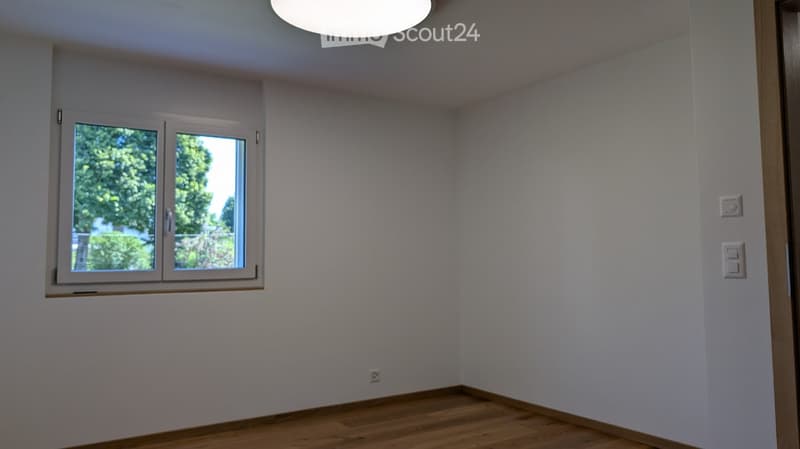 Empty room with light grey walls, wooden flooring, and a large window with white frame. The room has a modern ceiling light fixture and electrical outlets on the walls.