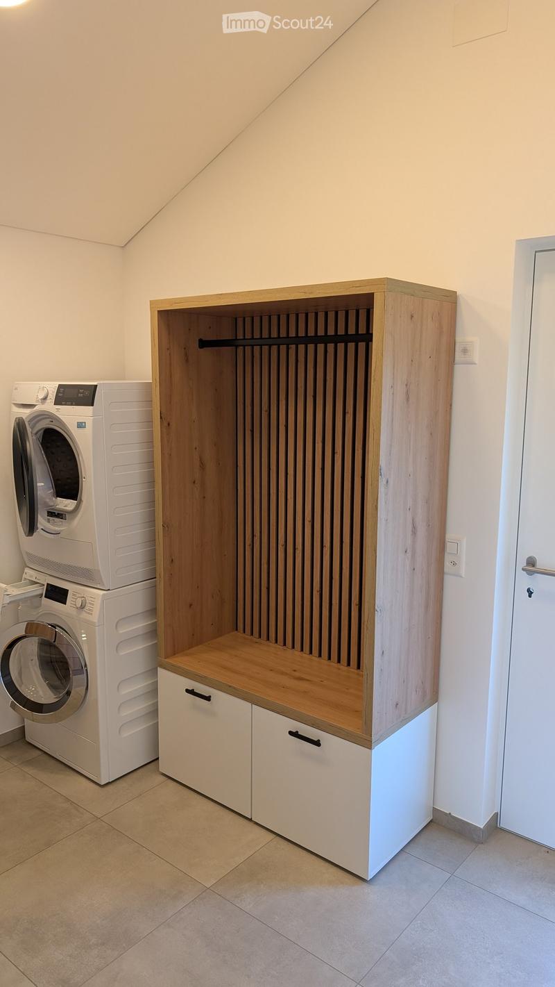 A small, well-lit room with a white washing machine and dryer stacked vertically, a wooden cabinet with shelves and drawers, and a tiled floor.