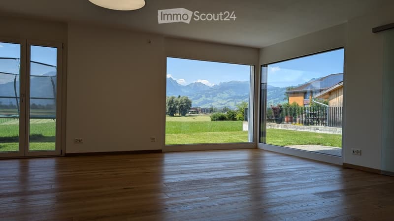 Empty room with wooden flooring, large windows offering a scenic mountain view, and modern lighting fixtures.