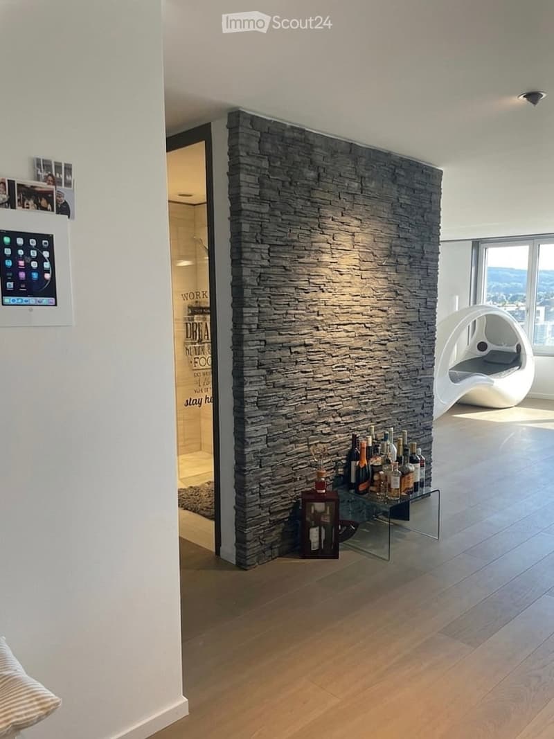 Open room with a stone wall, wooden floor, a glass table with bottles, a white chair, and a large window with a view