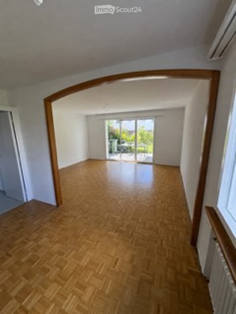 Empty room with wooden flooring, white walls, large window with glass door leading to a balcony or outdoor area, and a visible radiator on the wall.