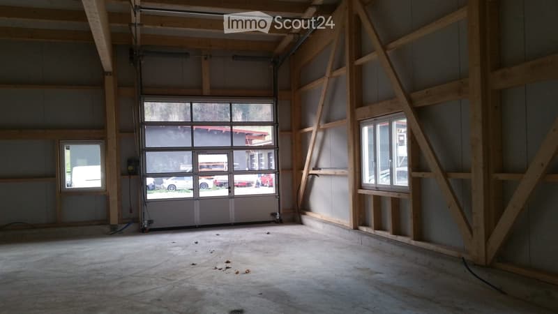 Empty garage with unfinished walls, exposed wooden beams, and a large garage door. The floor is concrete, and there are windows on the side walls.