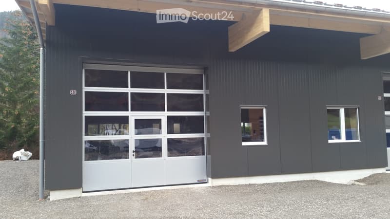 Modern single-story building with dark grey siding, large garage door with glass panels, and two small windows on the side.