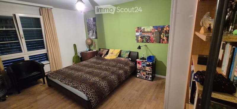 Cozy bedroom with a large bed, leopard print bedding, a black leather sofa, a nightstand with a lamp, a bookshelf, and a guitar leaning against the wall.