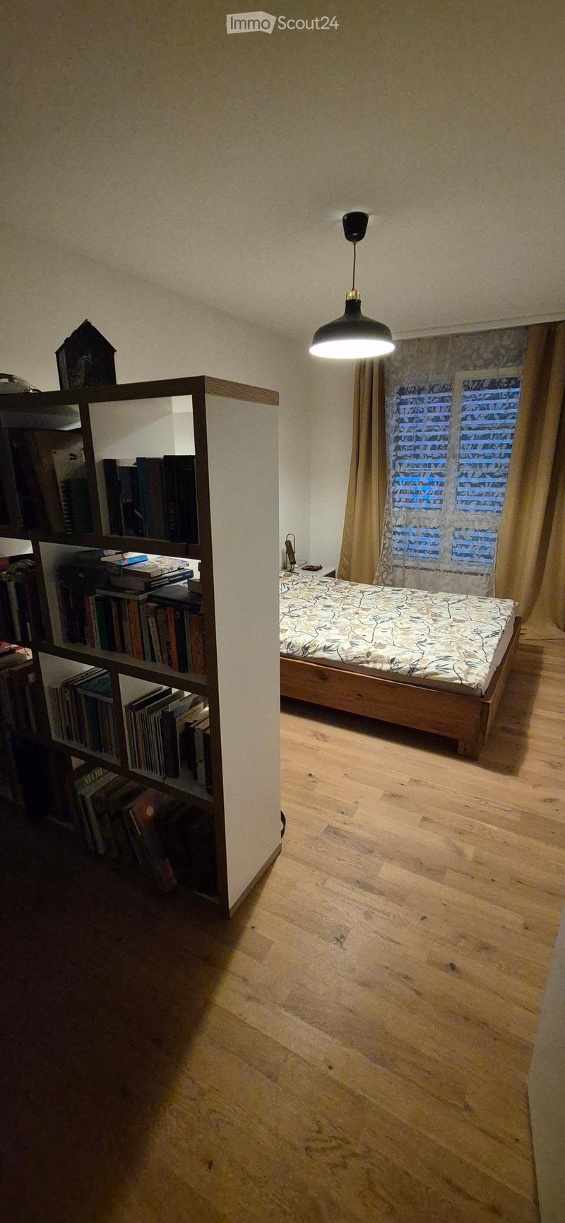Cozy bedroom with wooden floor, a bed with patterned bedding, a bookshelf filled with books, and a hanging lamp providing soft lighting.