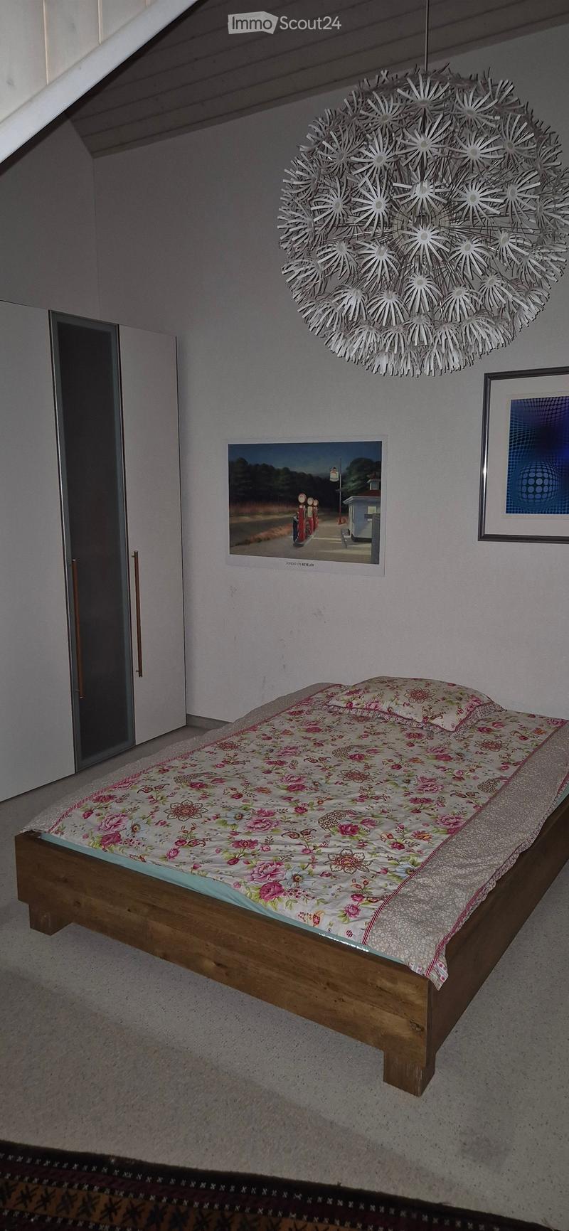 Bedroom with a wooden bed, floral bedspread, and a large decorative light fixture on the ceiling. There are two picture frames on the wall and a sliding glass door to the left.