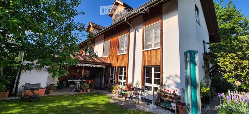 A well-maintained garden area with a lawn, patio furniture, potted plants, and a small shed. The house in the background has a mix of wooden and white exterior walls with multiple windows.