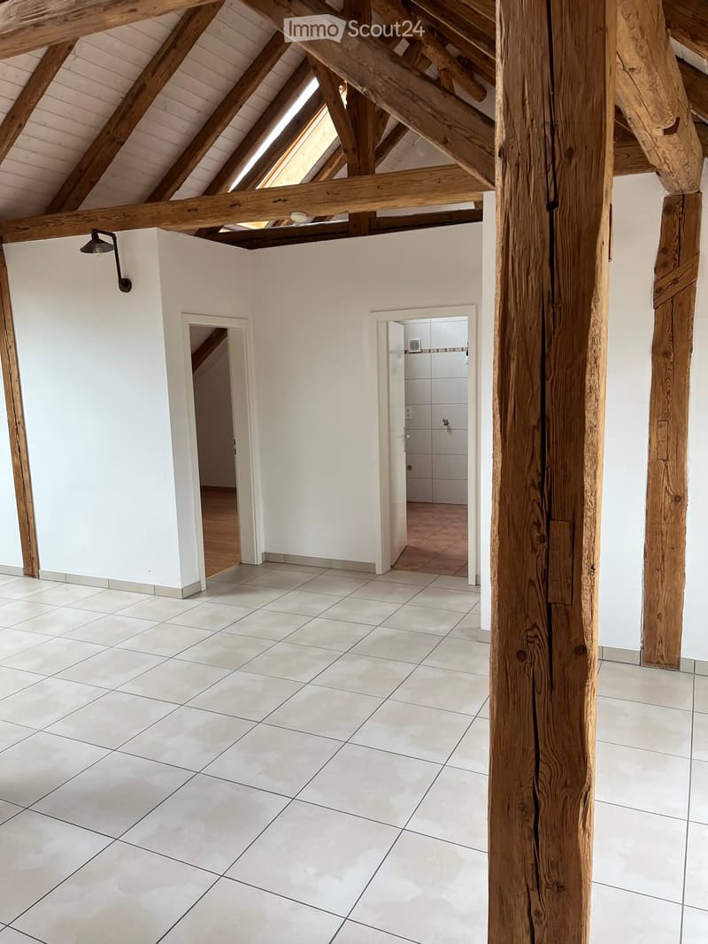 Bright, open room with white tiled flooring, exposed wooden beams, and white walls. Features a small hallway leading to another room and a partially visible bathroom with white tiles and a wooden door frame.