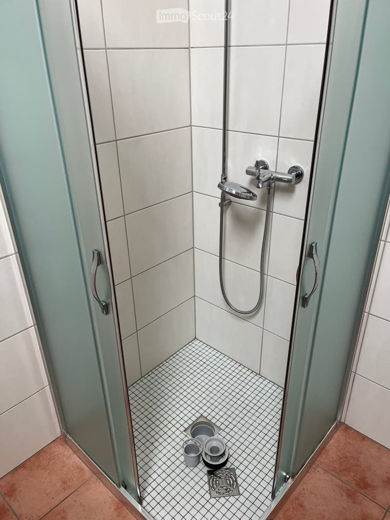 Modern shower stall with white tiles, glass enclosure, chrome fixtures, and a drain in the corner.