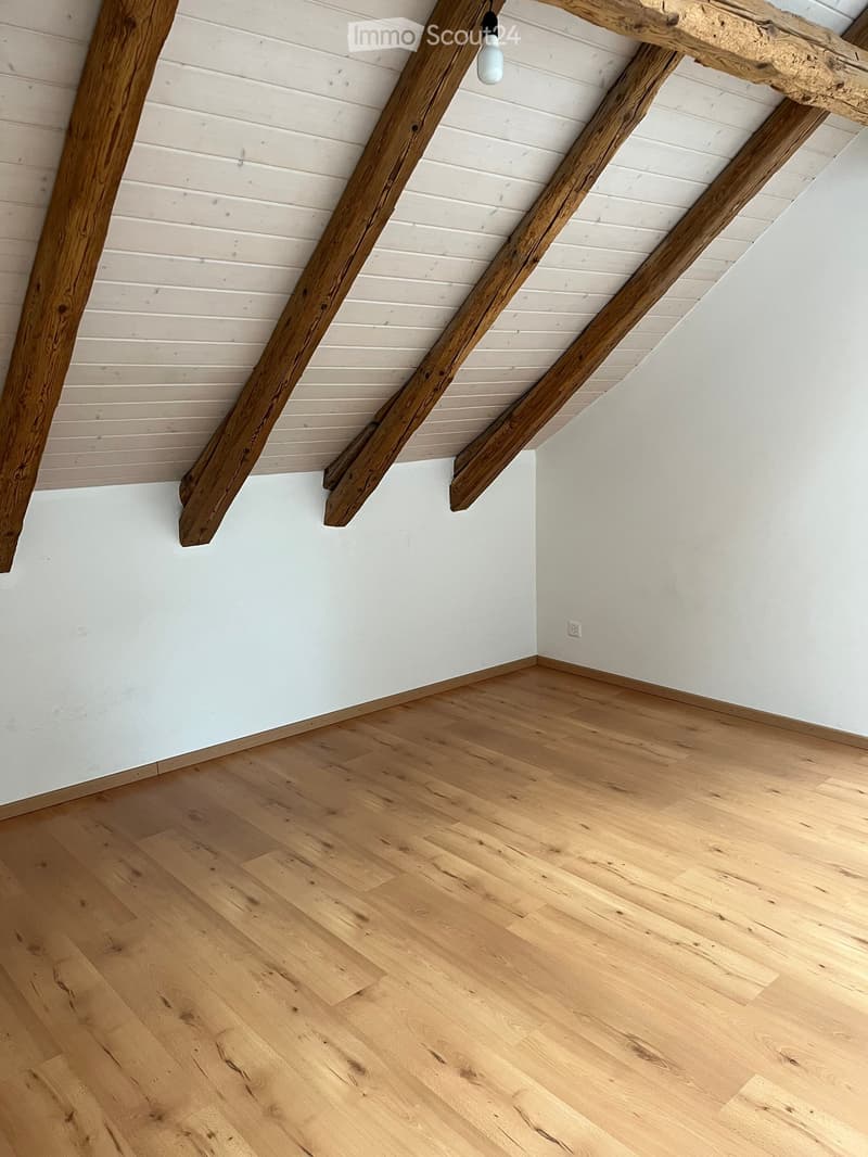 Empty room with light wooden flooring and white walls, featuring exposed wooden beams on the ceiling and a single hanging light fixture.