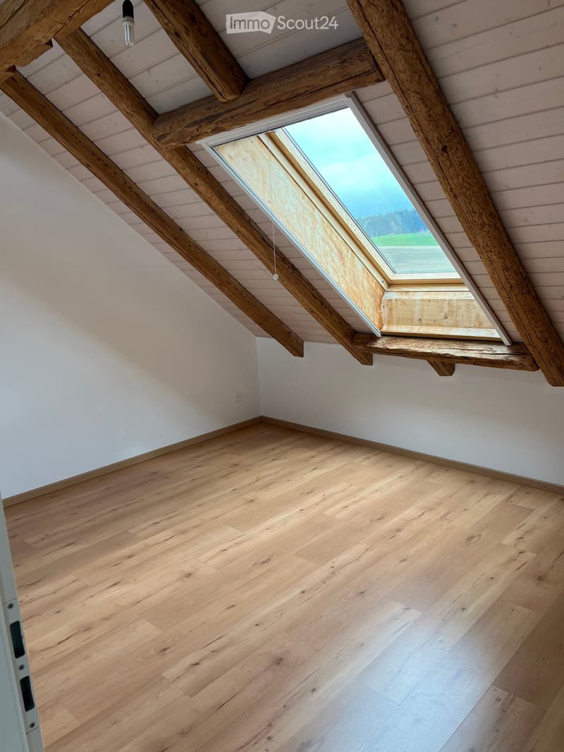Empty room with wooden flooring, white walls, and exposed wooden beams on the ceiling. A skylight window provides natural light.