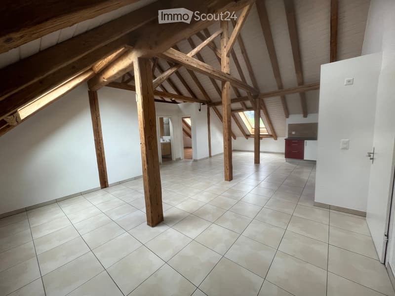 An empty room with exposed wooden beams and skylights, featuring tiled flooring and white walls. The space includes a small kitchen area with cabinets and a sink visible in the background.