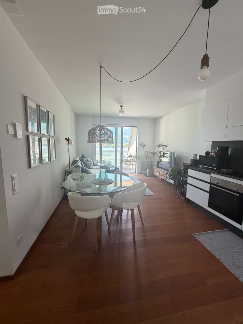 Modern open-plan living and dining area with wooden flooring, white furniture, and large windows leading to a balcony. Includes a glass dining table with white chairs, a sofa, kitchen area with cabinets and appliances, and decorative elements like picture