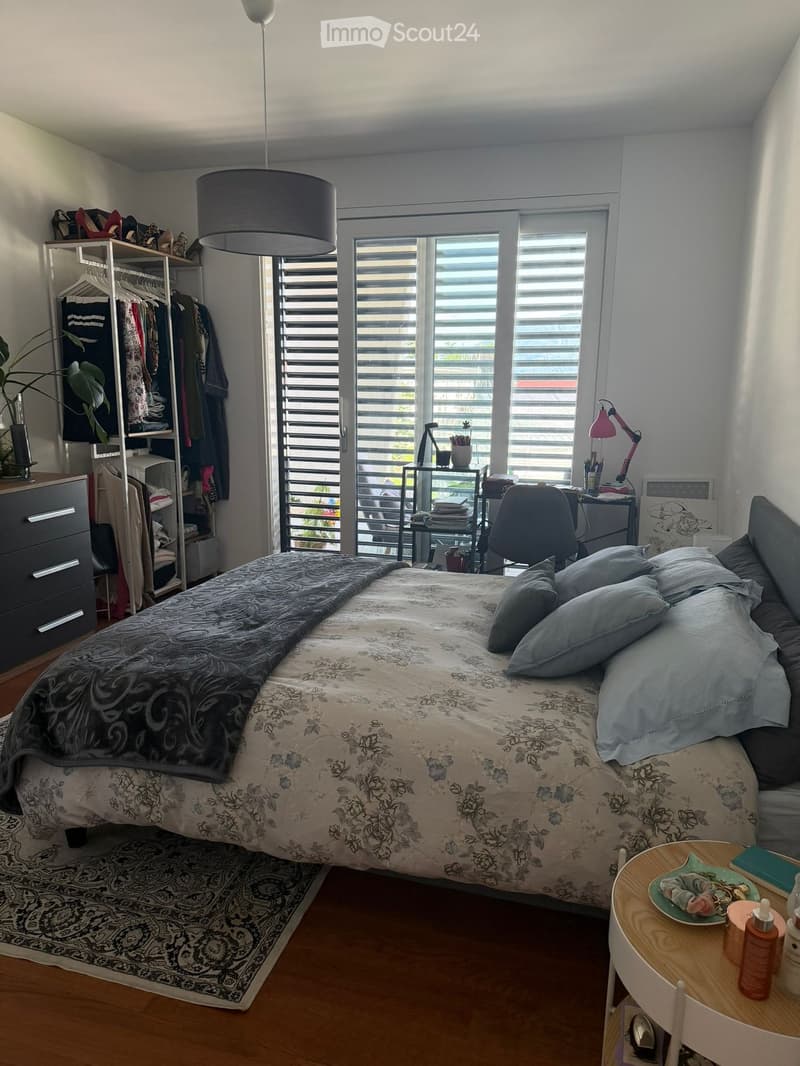 A well-lit bedroom with a double bed, nightstand, wardrobe, desk, and a small table with a chair. The room features large windows with blinds and a hanging lamp.