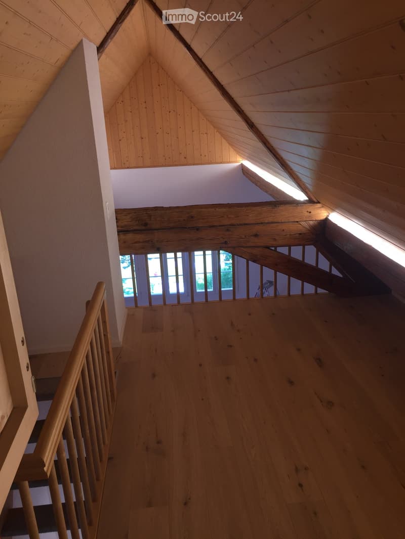 Wooden staircase with a slanted ceiling and skylight, leading to an open loft area with exposed wooden beams and a window.