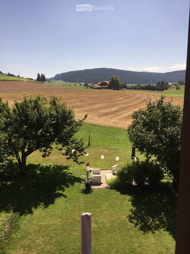 A well-maintained garden with a seating area, surrounded by trees and a scenic rural landscape with fields and hills in the background.