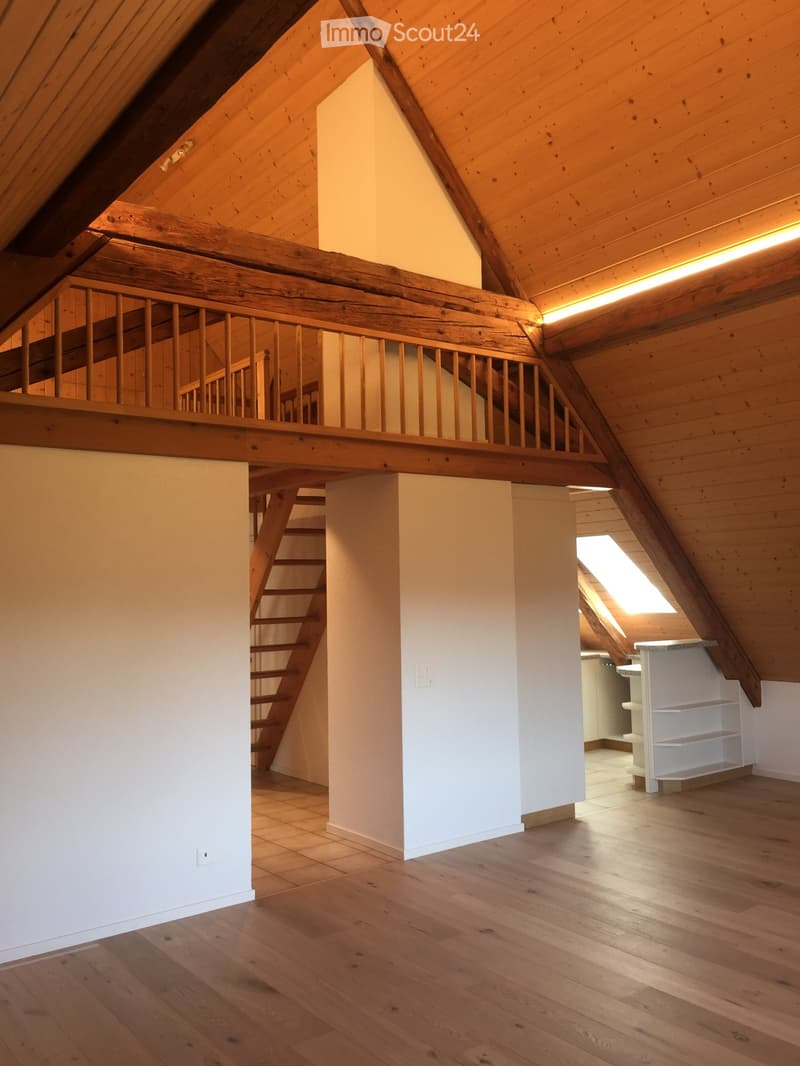 An attic space with wooden beams, exposed wooden ceiling, wooden floor, and a staircase leading to an upper level. The room is empty with white walls and a small window letting in natural light.