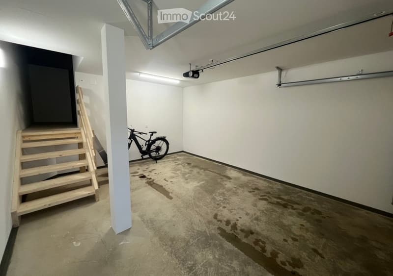Empty garage with concrete floor, white walls, wooden staircase, and a bicycle stored inside.