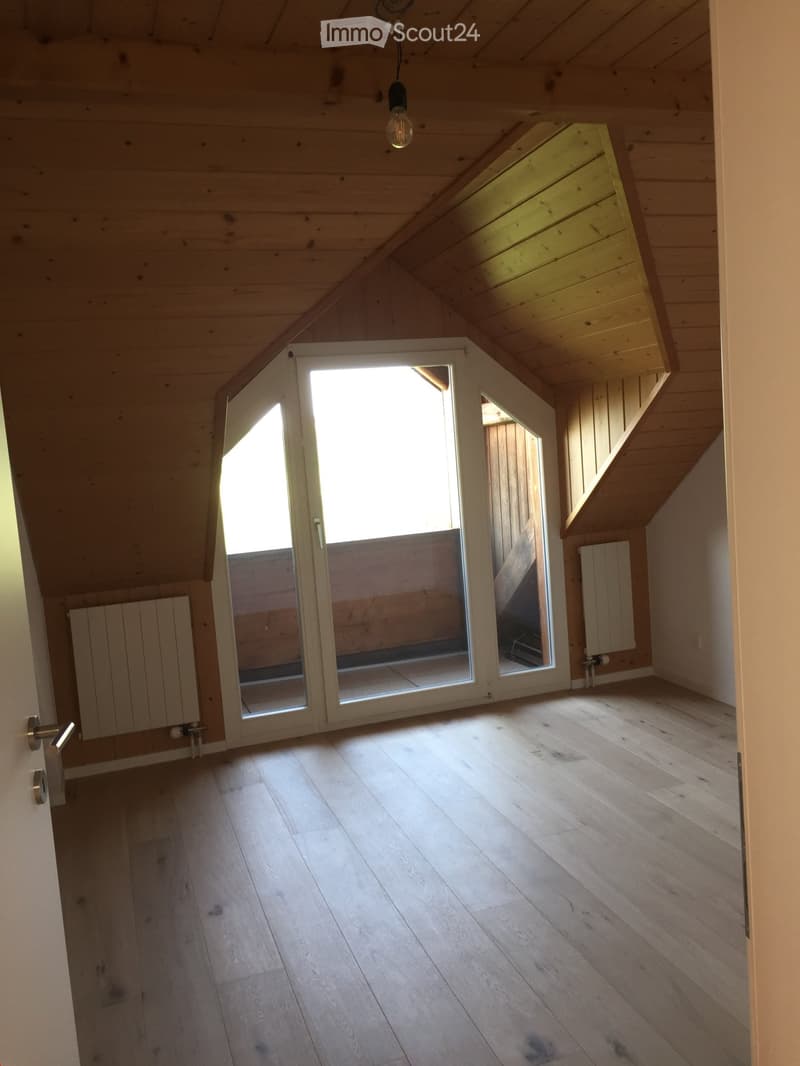 Empty attic room with wooden floors and slanted wooden ceilings, featuring large sliding glass doors that open to a balcony area.