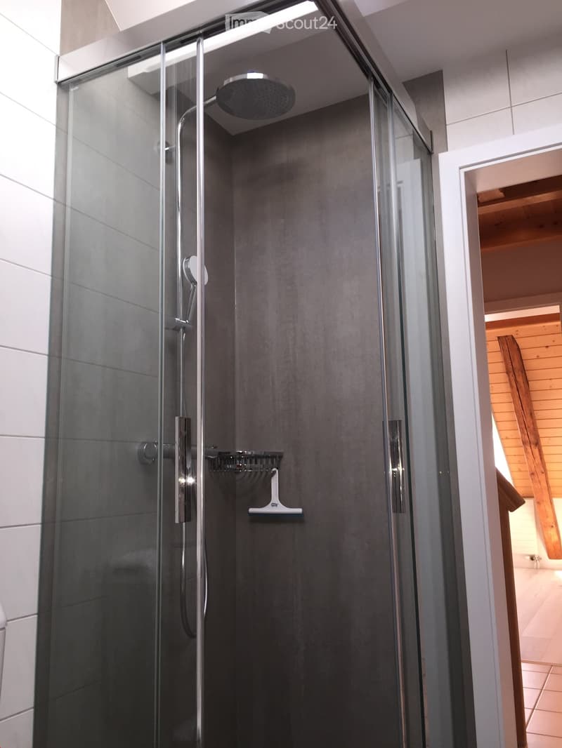 Modern shower area with a glass enclosure, dark grey tiles, and a handheld showerhead with a control handle. A small shelf is mounted inside the shower for storage.