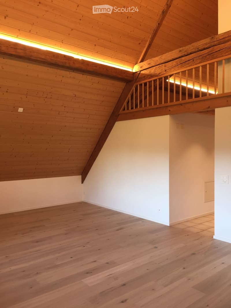 Empty room with wooden flooring and exposed wooden beams on the ceiling. The room features white walls and a wooden staircase railing leading to an upper level.