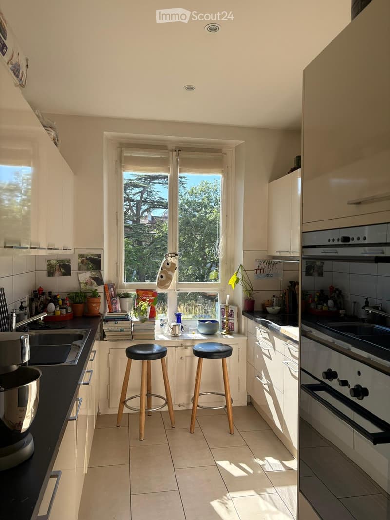 Bright kitchen with white cabinets, black countertops, and a large window overlooking greenery. Two wooden stools are placed in front of the island, and various kitchen items, including a sink, oven, and potted plants, are visible.