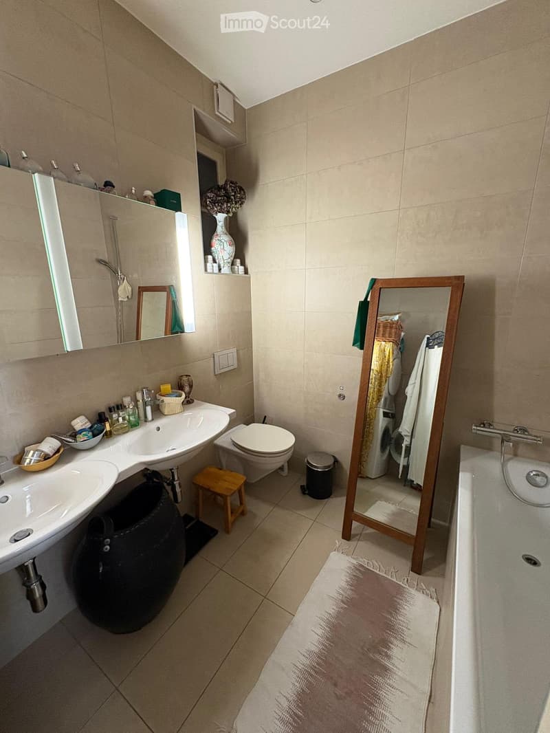 Modern bathroom with beige tiles, a double sink, toilet, bathtub, and a large mirror. Includes a small wooden stool, a decorative vase, and various toiletries.