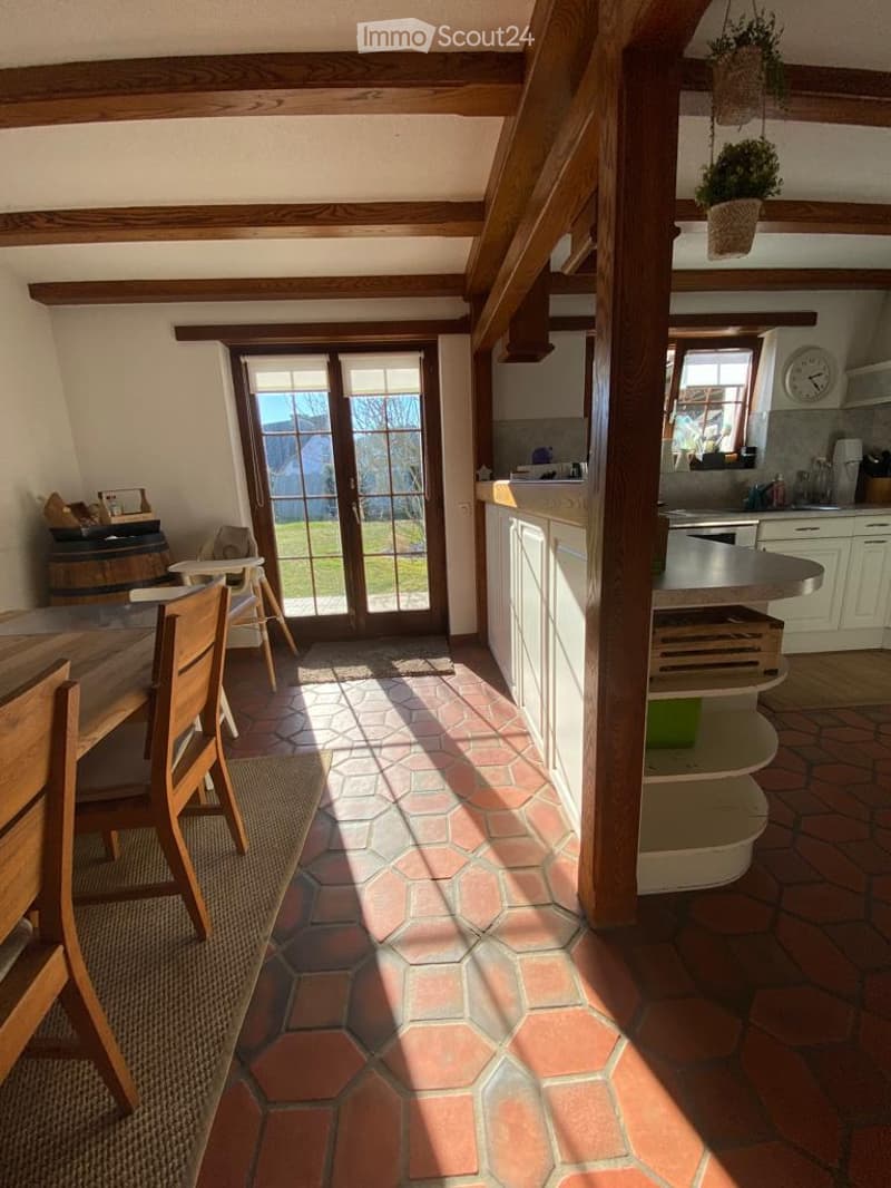Open-plan space with exposed wooden beams, tiled floor, large glass doors leading to outdoor area, dining table with chairs, bar counter, kitchen area with cabinets and sink.