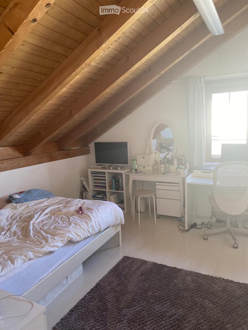 Cozy attic bedroom with wooden slanted ceilings, a neatly made bed, a desk with a laptop, a mirror, and a small rug on the floor.