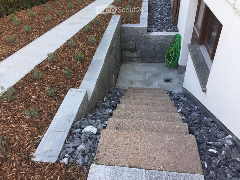 A raised garden pathway leading to a house entrance, bordered by concrete walls and mulch beds with small plants.