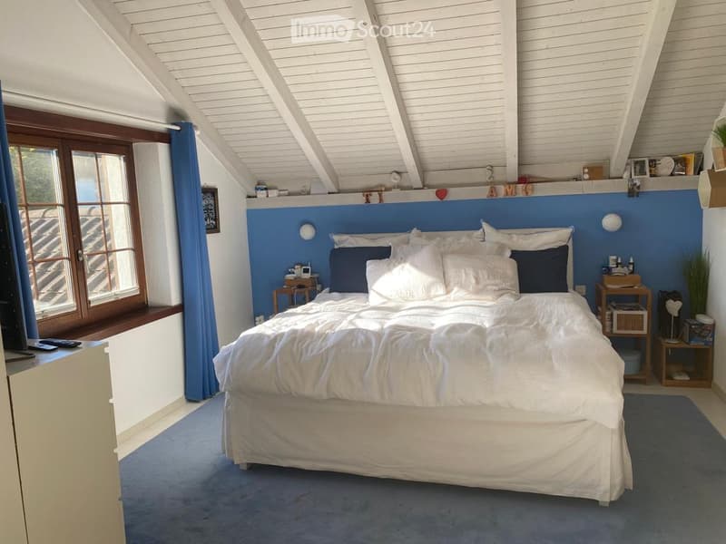 Bright bedroom with a large window offering a view, featuring a neatly made bed with white and blue bedding, nightstands, a small table, and a chair.