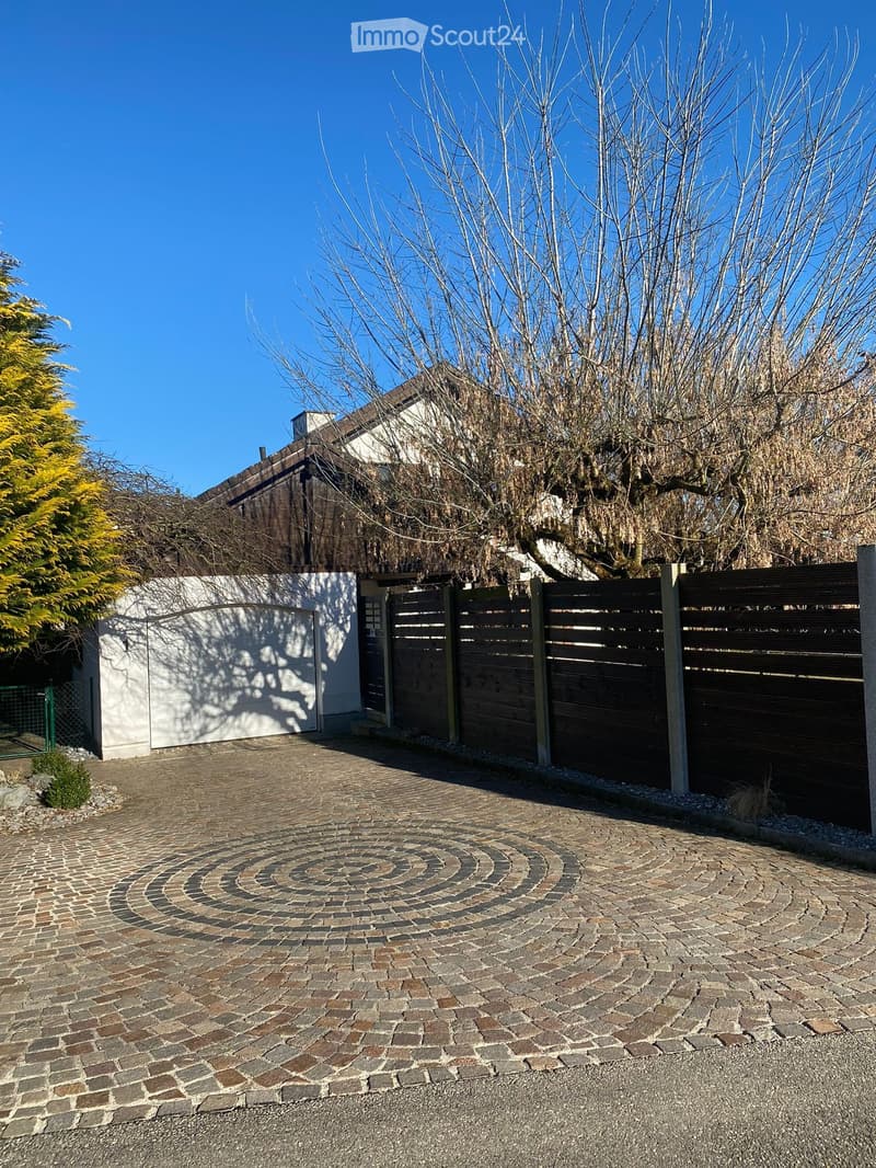A driveway with a spiral brick pattern leads to a house entrance, surrounded by a wooden fence and bare trees.