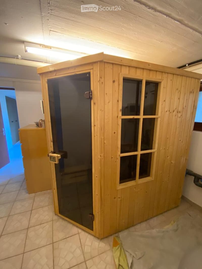 A wooden sauna cabin with glass door and windows, placed inside a room with tiled flooring and white walls.