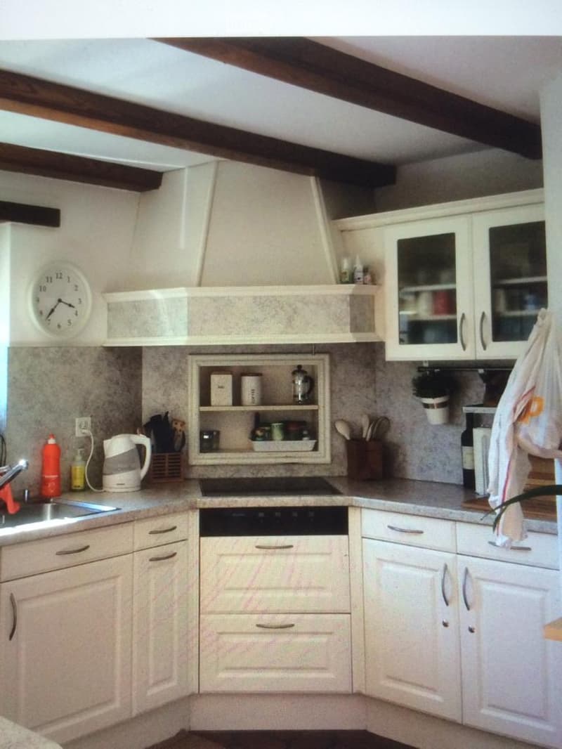A modern kitchen with white cabinets, a central island, and various appliances including a dishwasher, oven, and sink. The space features a tiled backsplash, wooden beams on the ceiling, and a clock on the wall.