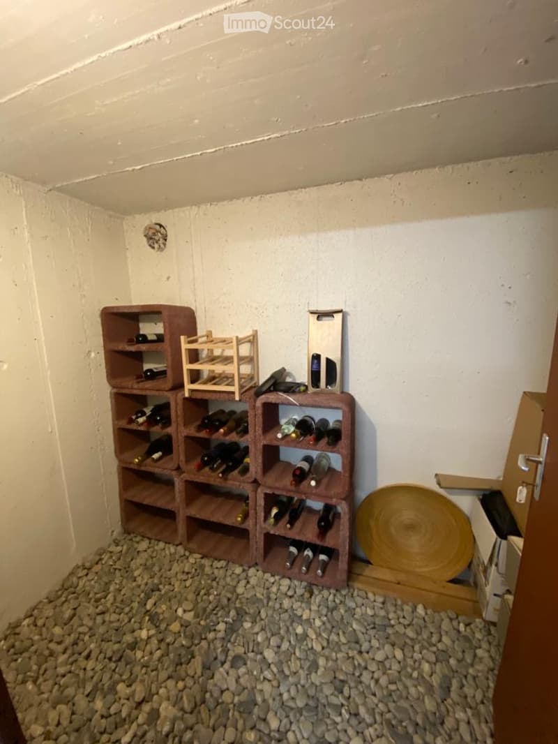 A small, cozy wine cellar with wooden racks and shelves holding multiple wine bottles, a round wooden disc on the floor, and a wooden crate on top of the racks.