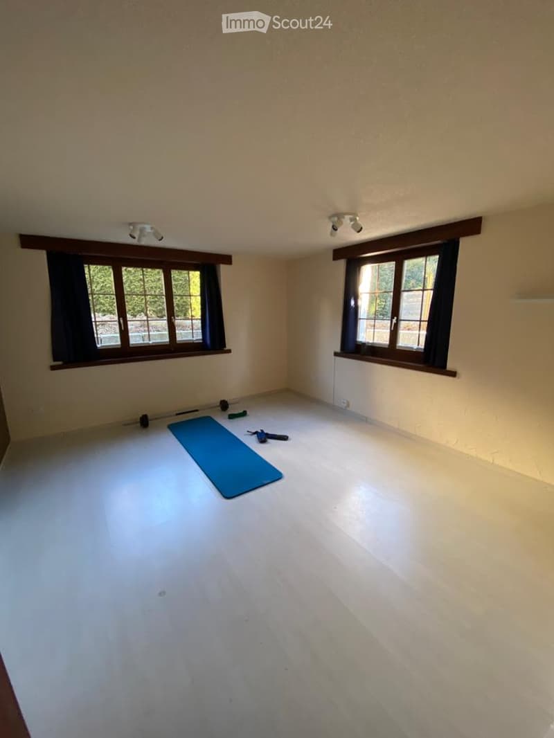 Empty room with light-colored walls, wooden floor, and large windows with dark curtains. A blue exercise mat and a few gym accessories are placed on the floor.