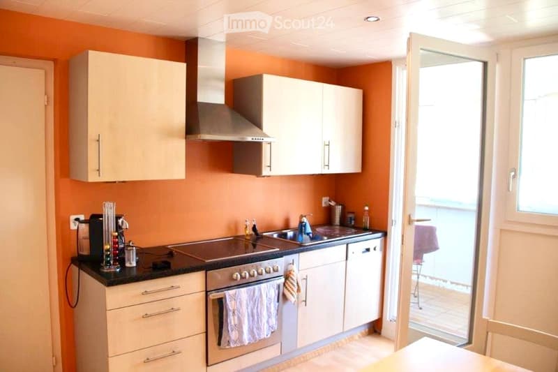 Modern kitchen with light-colored cabinets, stainless steel appliances, and a black countertop. An open door leads to a balcony.