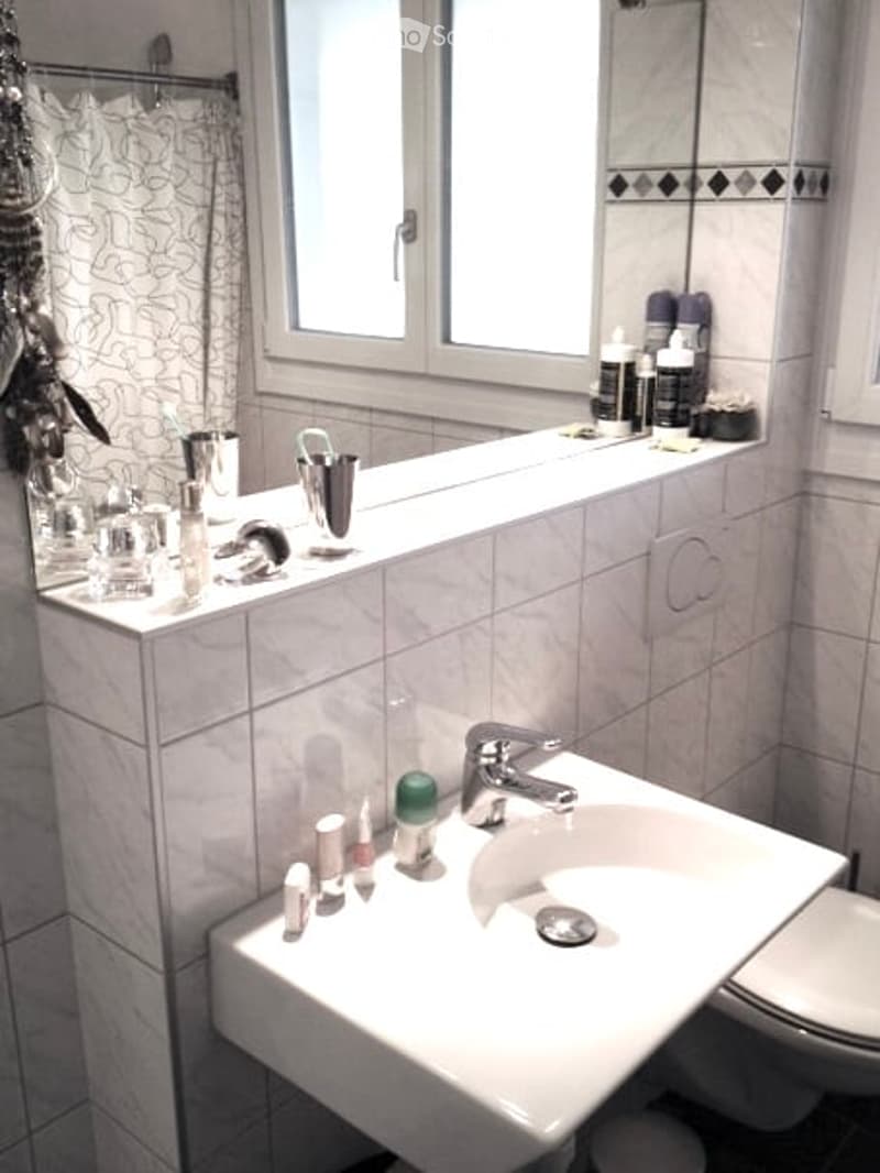 Modern bathroom with white tiles, a large window, a sink with a faucet, a toilet, a mirror, and various toiletries on the counter.