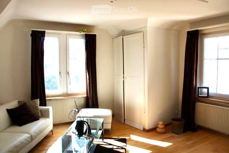 Living room with light-colored sofa, coffee table, ottoman, and a small screen on a windowsill. The room has wooden flooring, large windows with dark curtains, and a small plant on the coffee table.