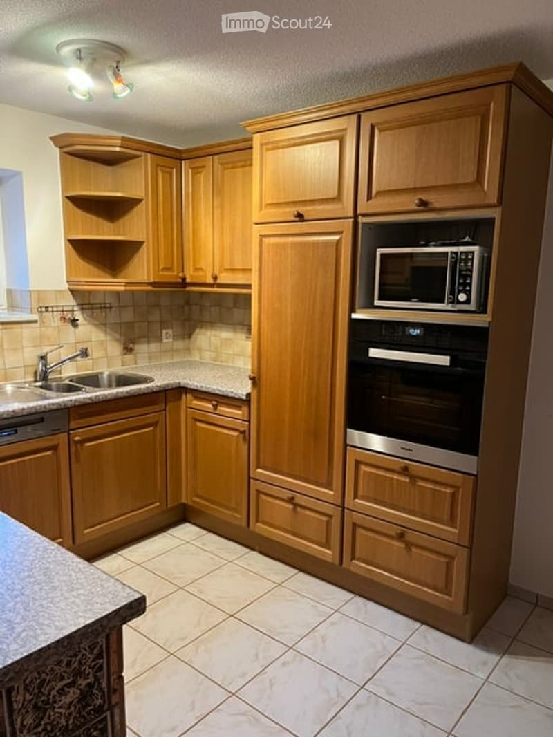 A modern kitchen with wooden cabinets, a stainless steel sink, microwave, and oven. The countertops are granite, and the floor is tiled.