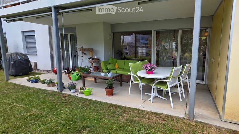 A covered outdoor patio area with a green sofa, a white dining table with chairs, potted plants, and a small garden area with grass and additional plants.