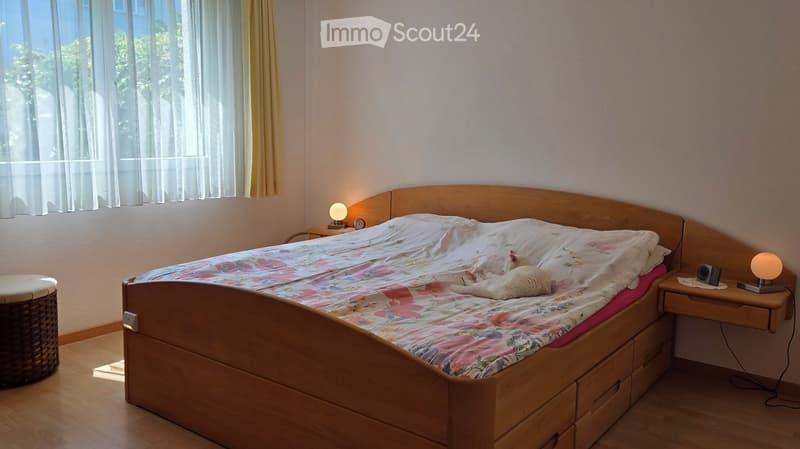 Cozy bedroom with a wooden bed, floral bedding, a small nightstand with a lamp, and a wicker stool near the window.