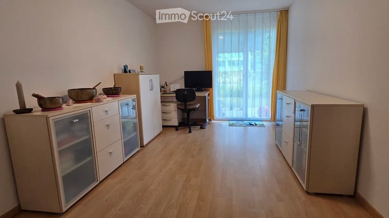 A furnished room with a wooden floor, a refrigerator, a desk with a screen, a chair, and a small cabinet with a bowl on top.
